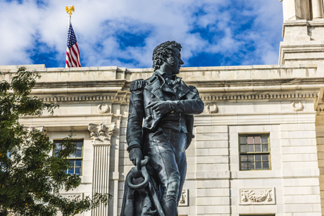 Bronze statue in front of a neoclassical building with an American flag and blue sky