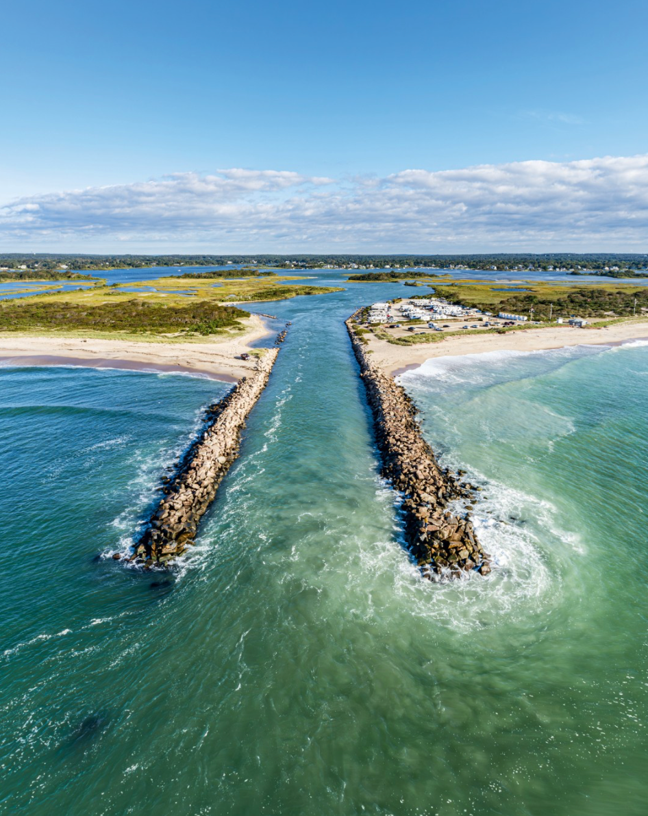 Aerial view of a narrow inlet between sandy beaches and rock jetties, with blue-green water and a distant coastline