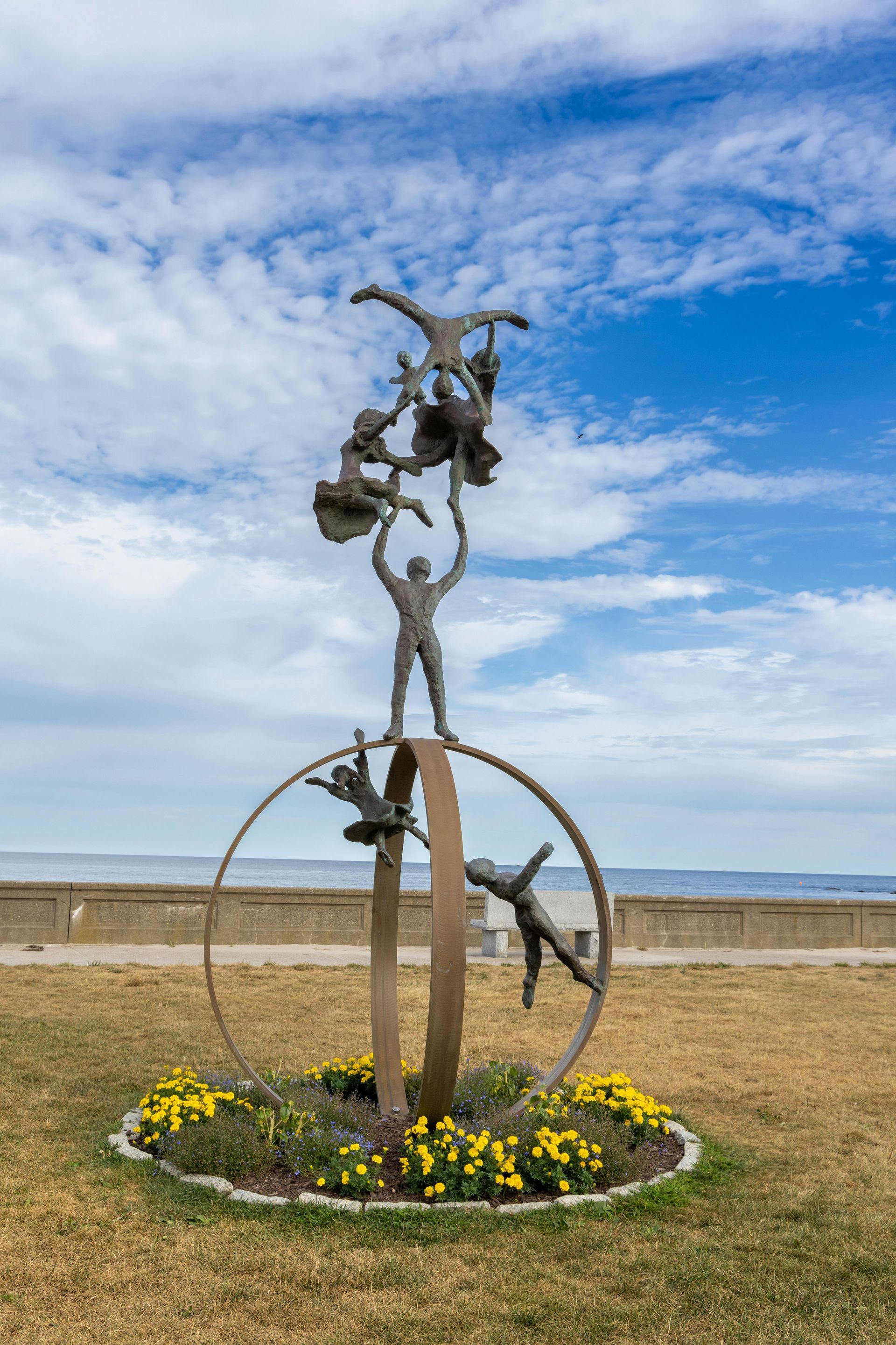 Metal sculpture in a flower bed by a beach, with blue sky and ocean in the background