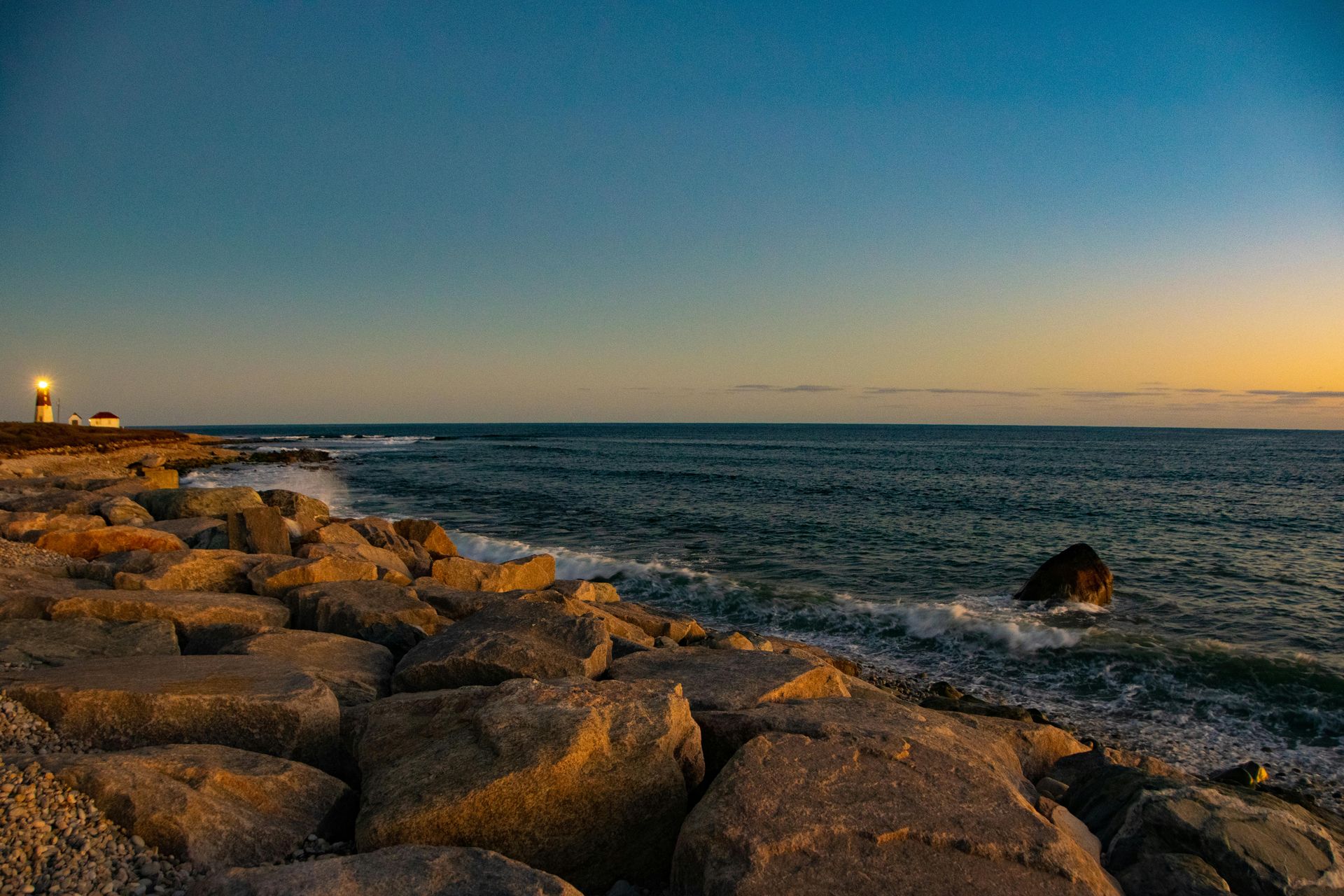 Rocky shoreline at sunset with calm ocean, glowing horizon, and a distant lighthouse on the left