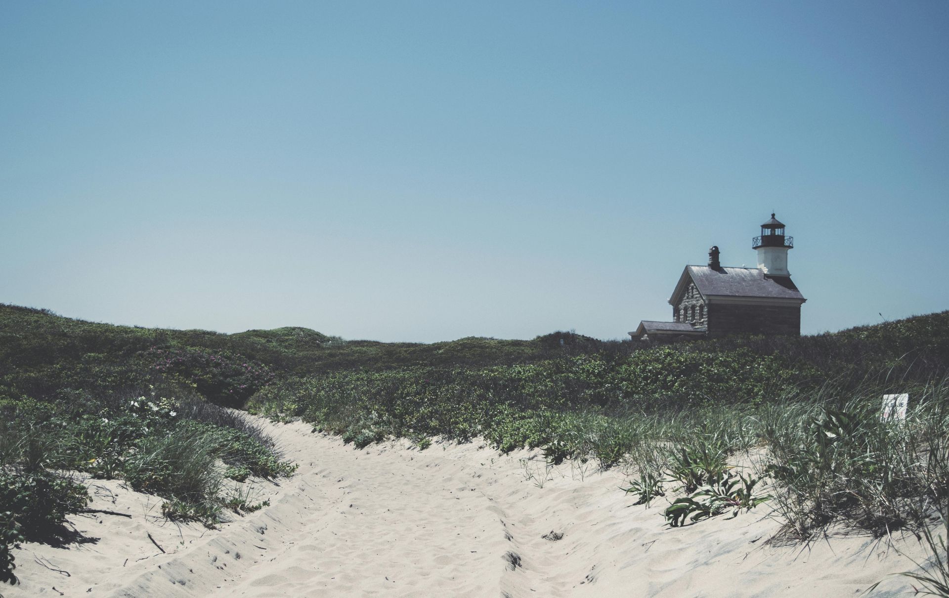 Sandy path through dunes leading to a white lighthouse under a clear blue sky