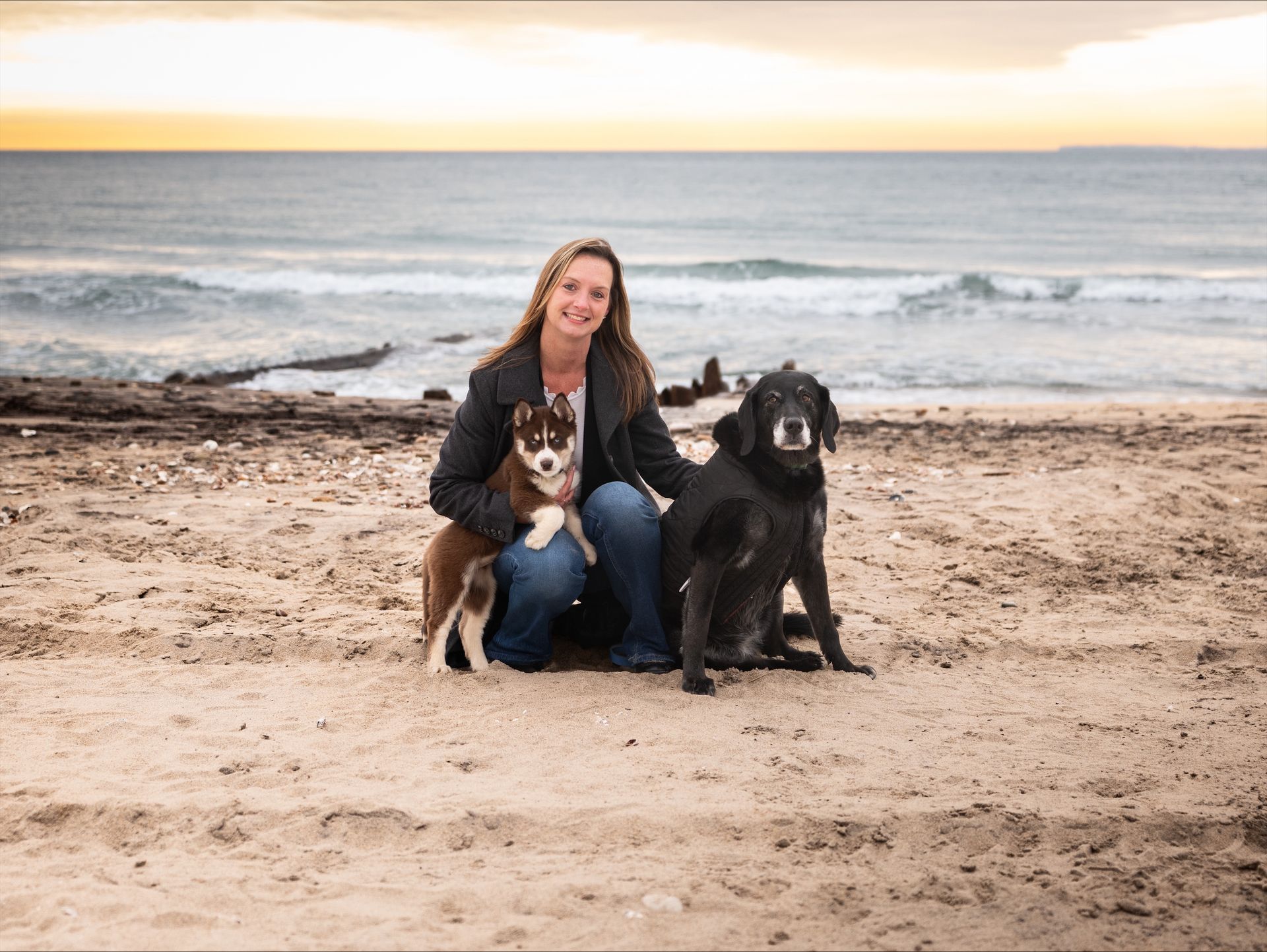 Woman kneeling on a beach with two dogs, ocean waves and a sunset sky behind them