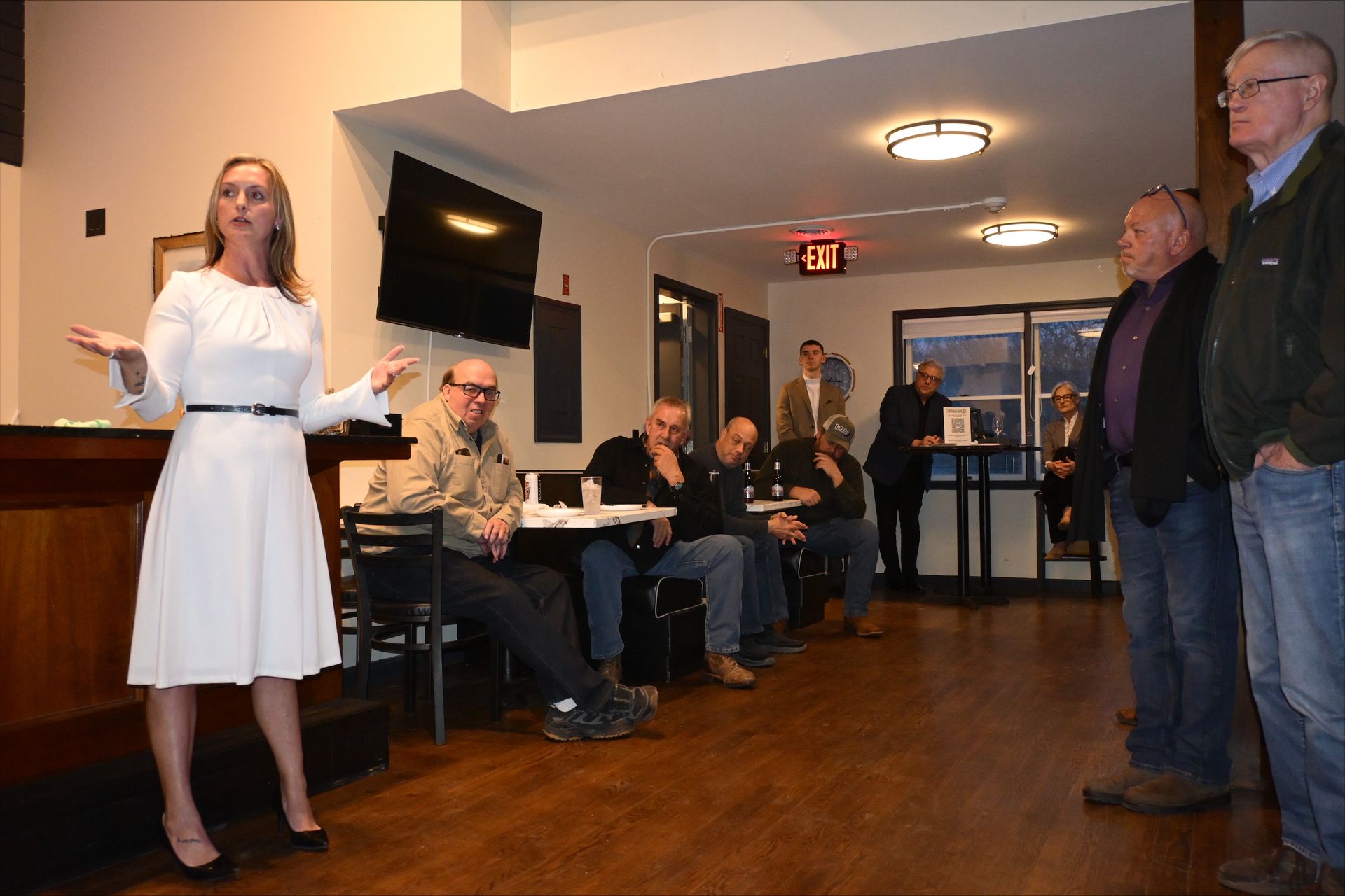 Woman speaking to a small group in a hallway, with seated and standing attendees listening.