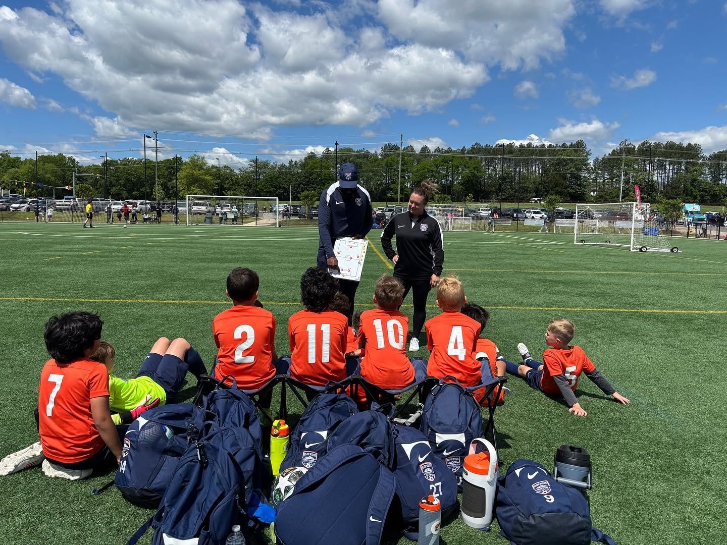 Youth soccer team listening to coaches on a sunny field, orange jerseys and blue bags in the foreground