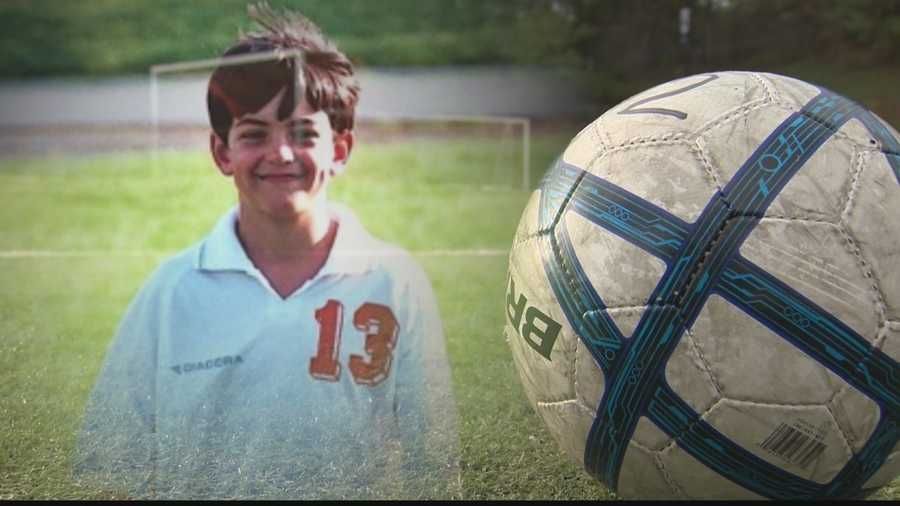 Boy in a white soccer jersey stands on a field beside a close-up soccer ball.