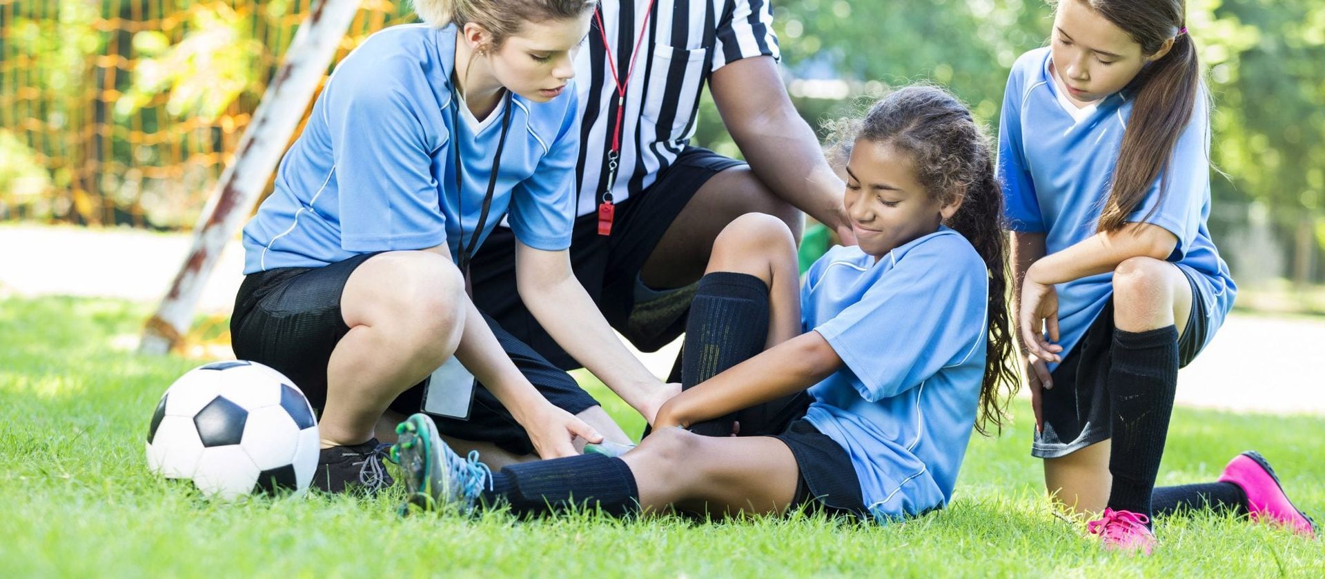 Children and a coach sitting on grass during a soccer game, helping an injured child with a soccer ball nearby