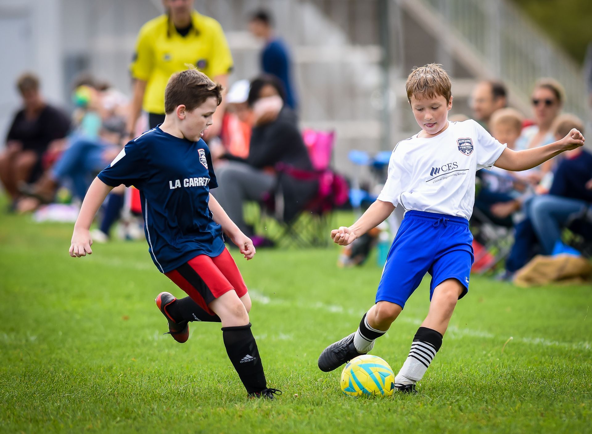 Two children playing soccer on a grassy field, one in navy and red, the other in white and blue