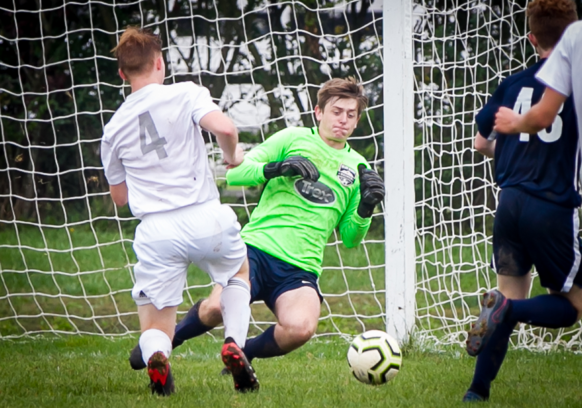 Soccer goalkeeper in green blocks a shot near the goal as two players in white and navy close in