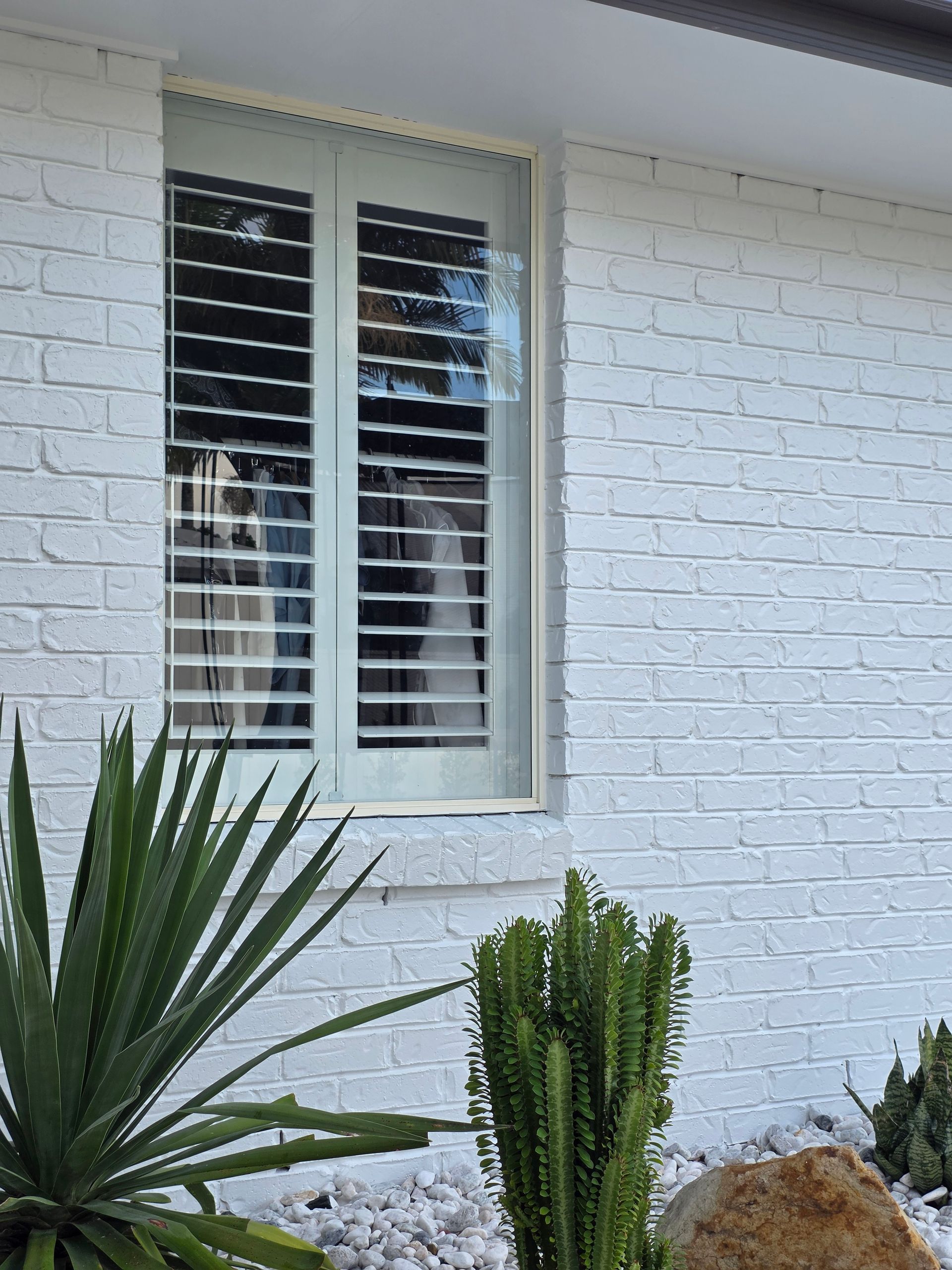 White brick house wall with a shuttered window and green plants in front.