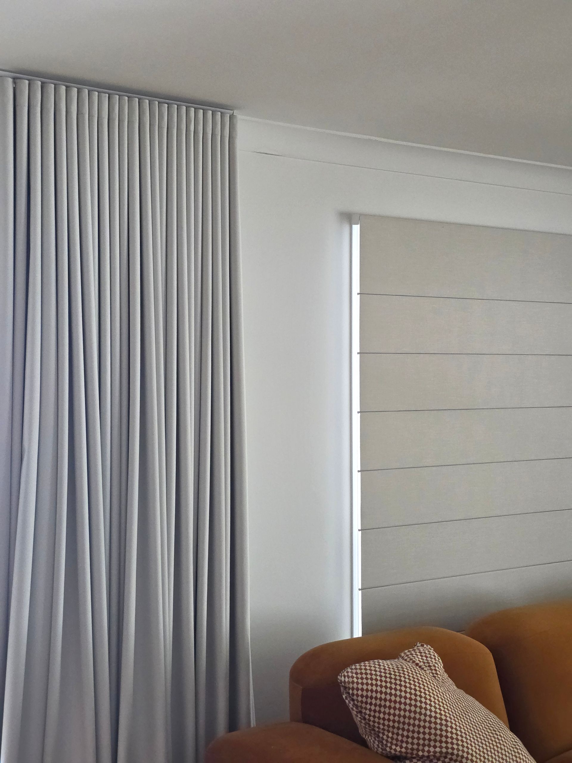 Modern living room with gray curtains, a window blind, and part of an orange sofa with a patterned pillow