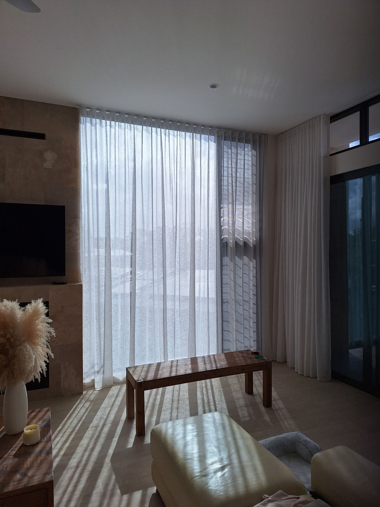 Bright living room with sheer curtains, wooden bench, armchair, and sunlight near a glass door.