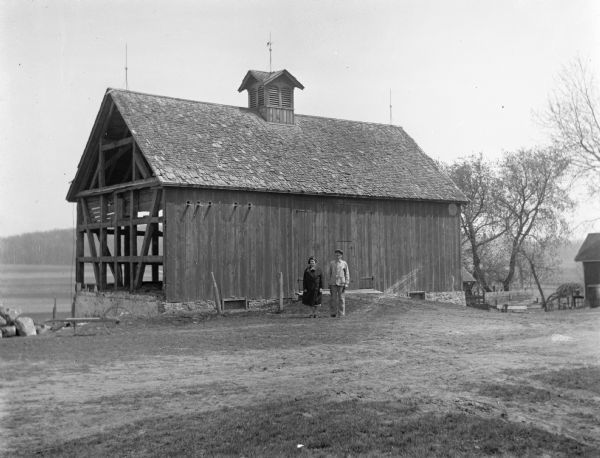 A black and white photo of a large rustic wooden barn, set near a body of water and surrounded by a rural landscape, representing barns of America.