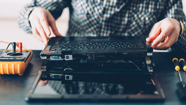 Hands in a plaid shirt carefully disassemble or repair an open laptop on a dark desk with a multimeter and screwdrivers.