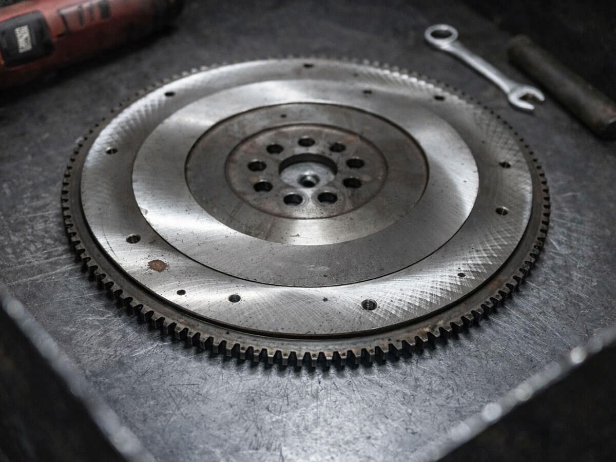 A steel flywheel for a vehicle engine sits on a metallic workshop surface alongside a wrench and a tool handle.
