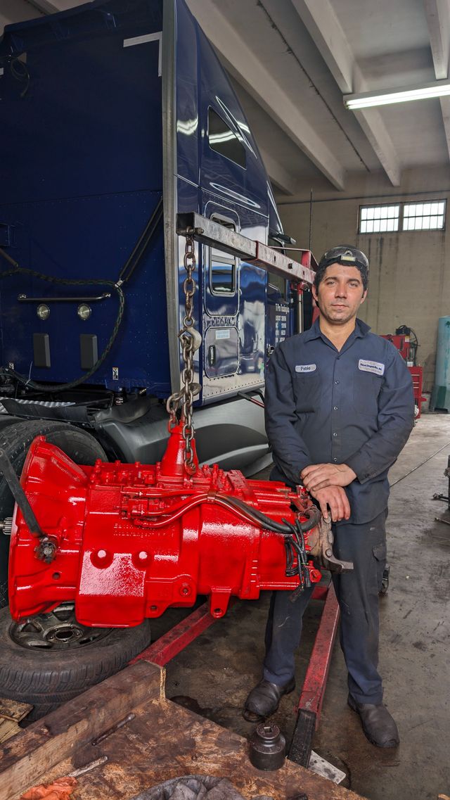 A mechanic standing next to a bright red truck transmission in a repair shop with a large blue truck in the background.