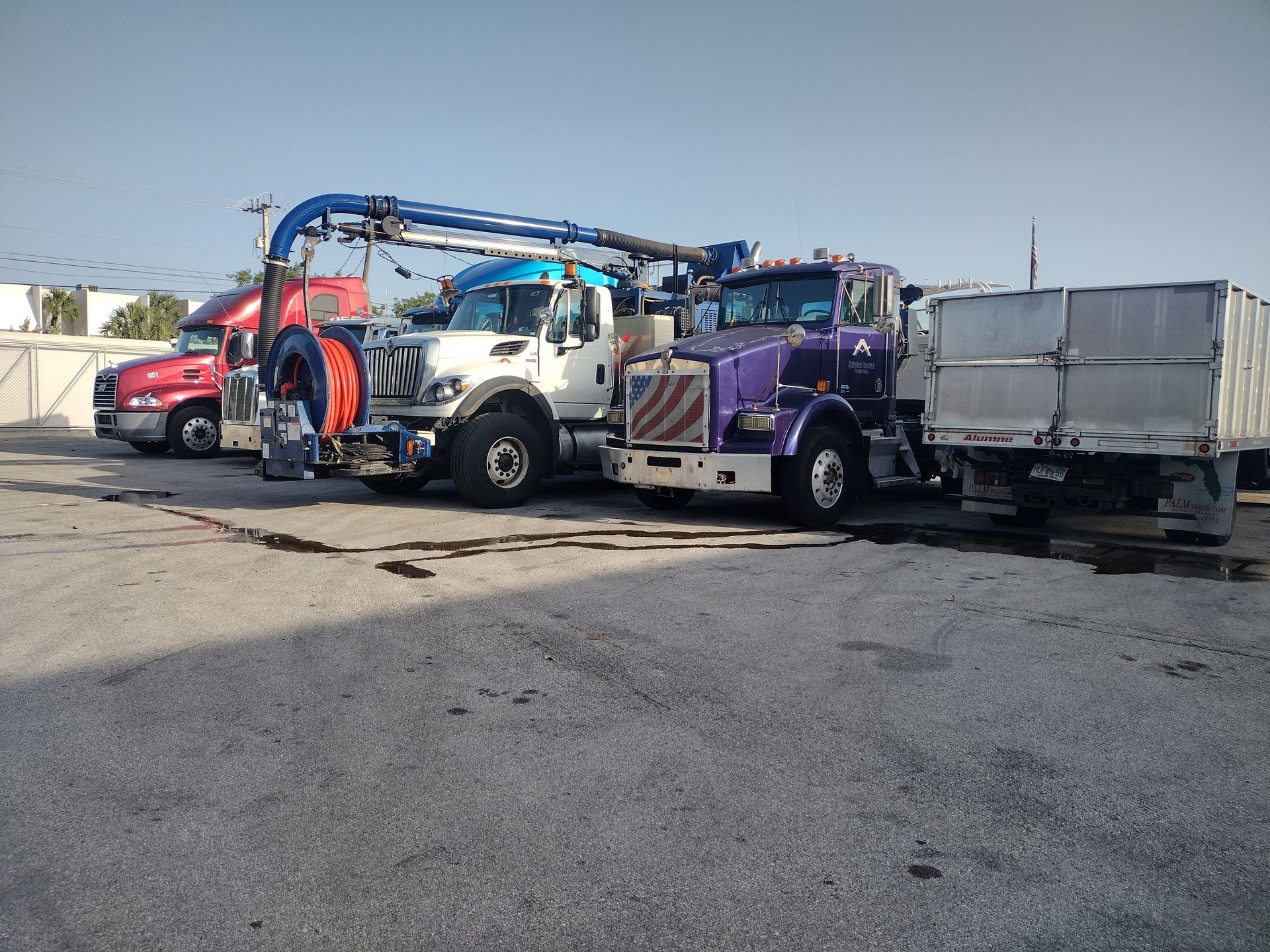 Three industrial trucks, including a white and purple vacuum truck with a boom arm, parked in a paved lot.