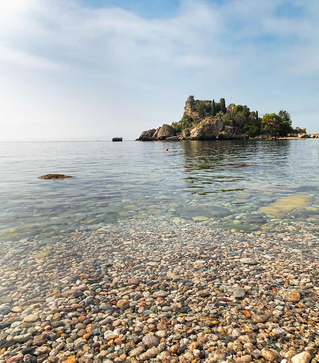 Vista costiera con acqua limpida su una riva di ciottoli, che conduce a una piccola isola sotto un cielo azzurro.