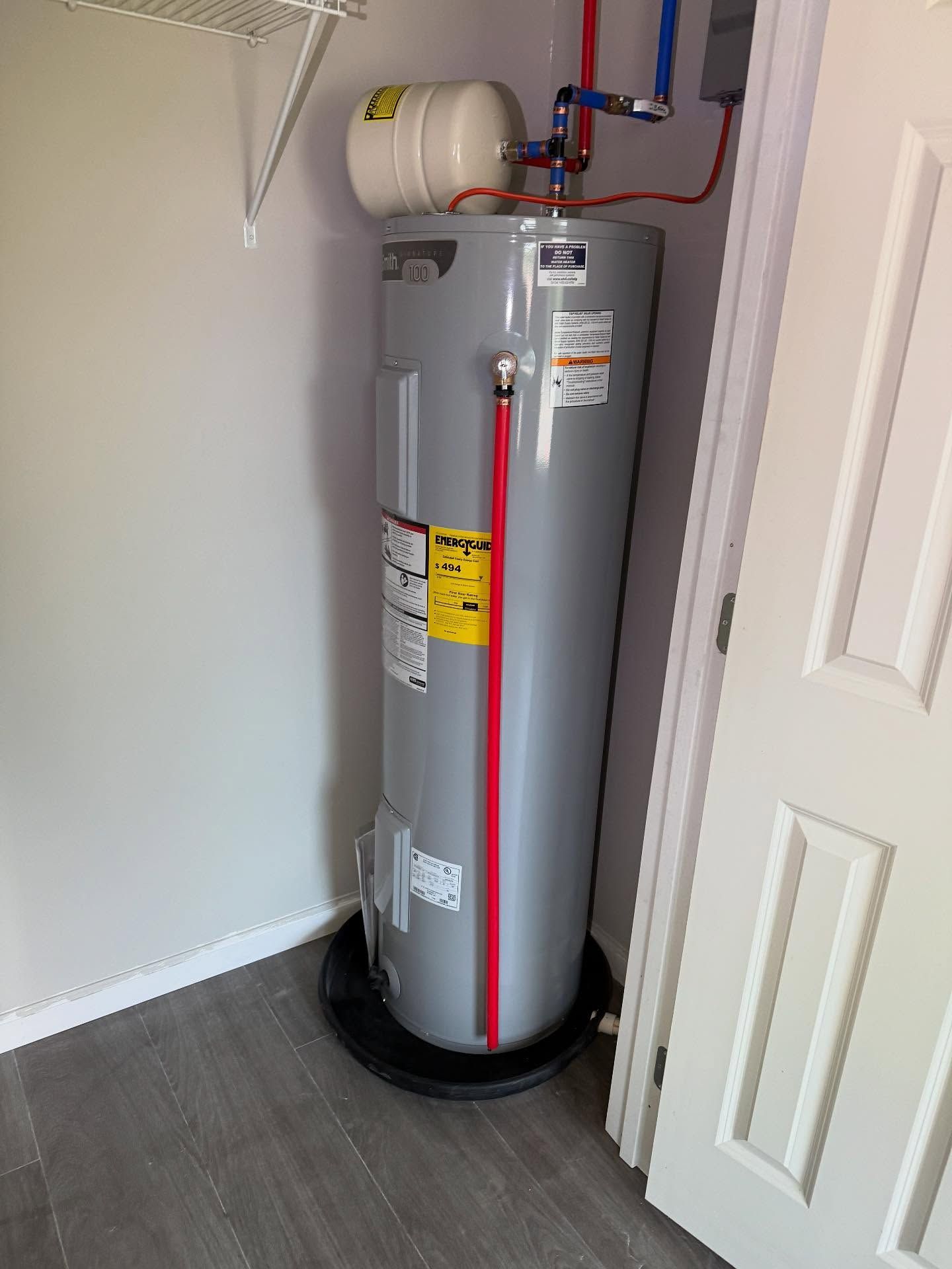 Tall gray water heater in a closet with red and blue pipes and a white expansion tank.