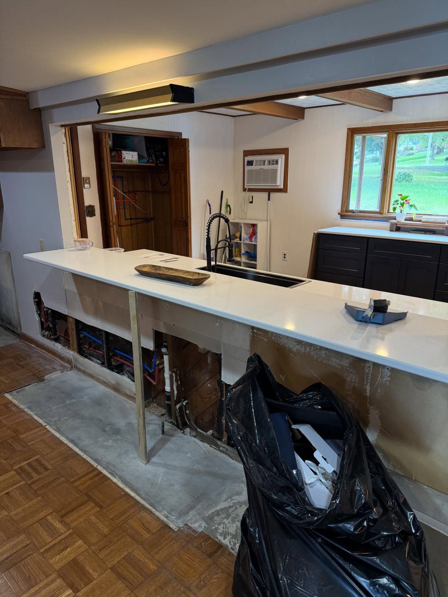 Unfinished kitchen bar with white countertop, exposed framing, cabinets, and a window view.