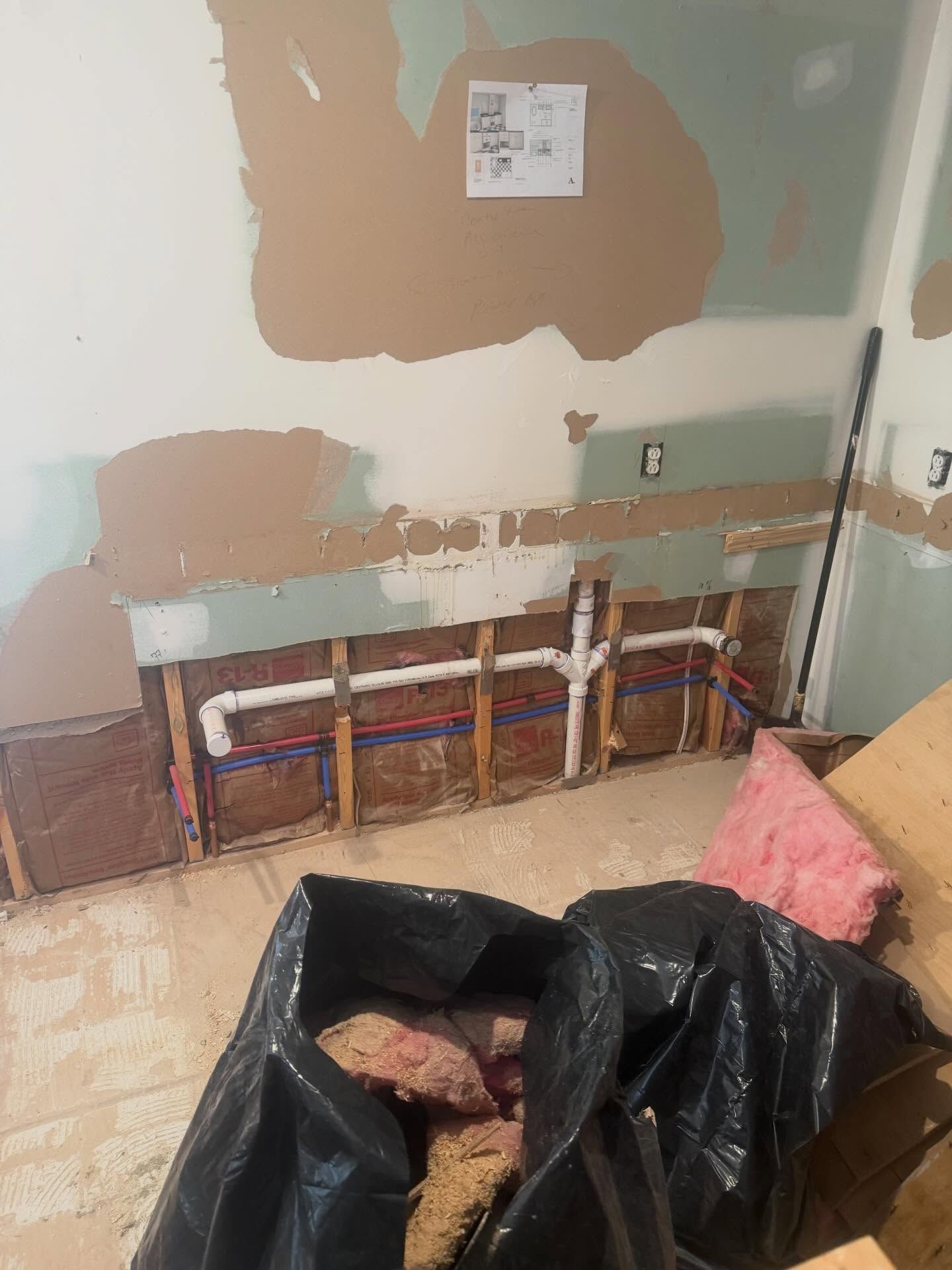 Partially demolished room wall with exposed plumbing, black trash bags, and debris on the floor