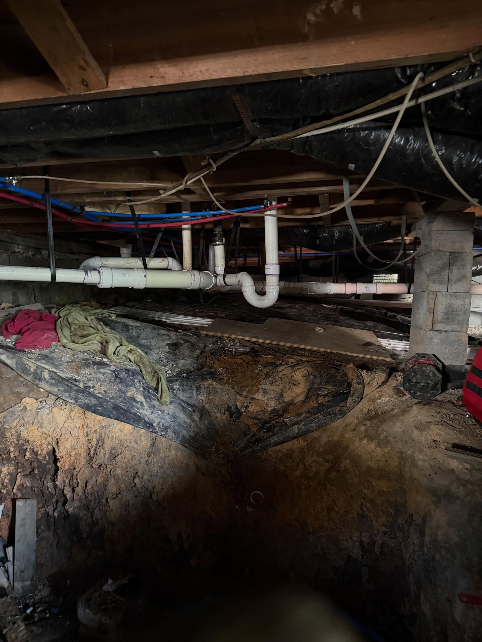 Exposed basement plumbing and pipes beneath floor joists over an earthen crawlspace.