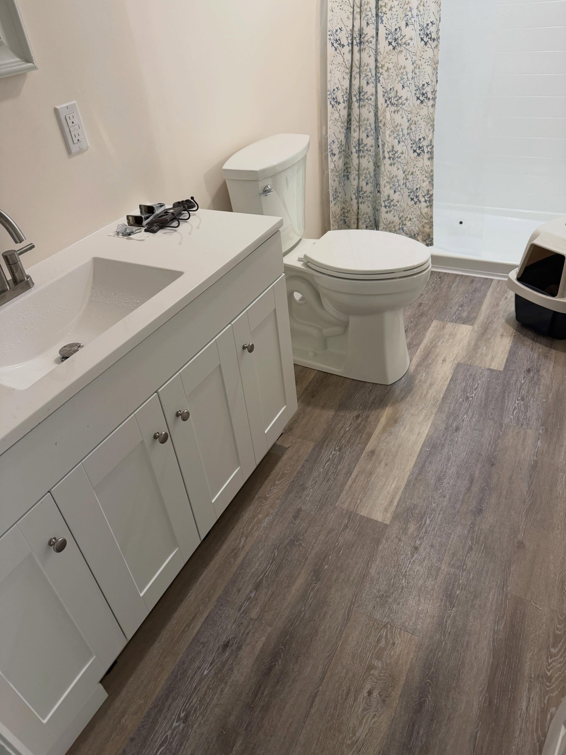 Small bathroom with white vanity, toilet, floral shower curtain, and wood-look flooring.
