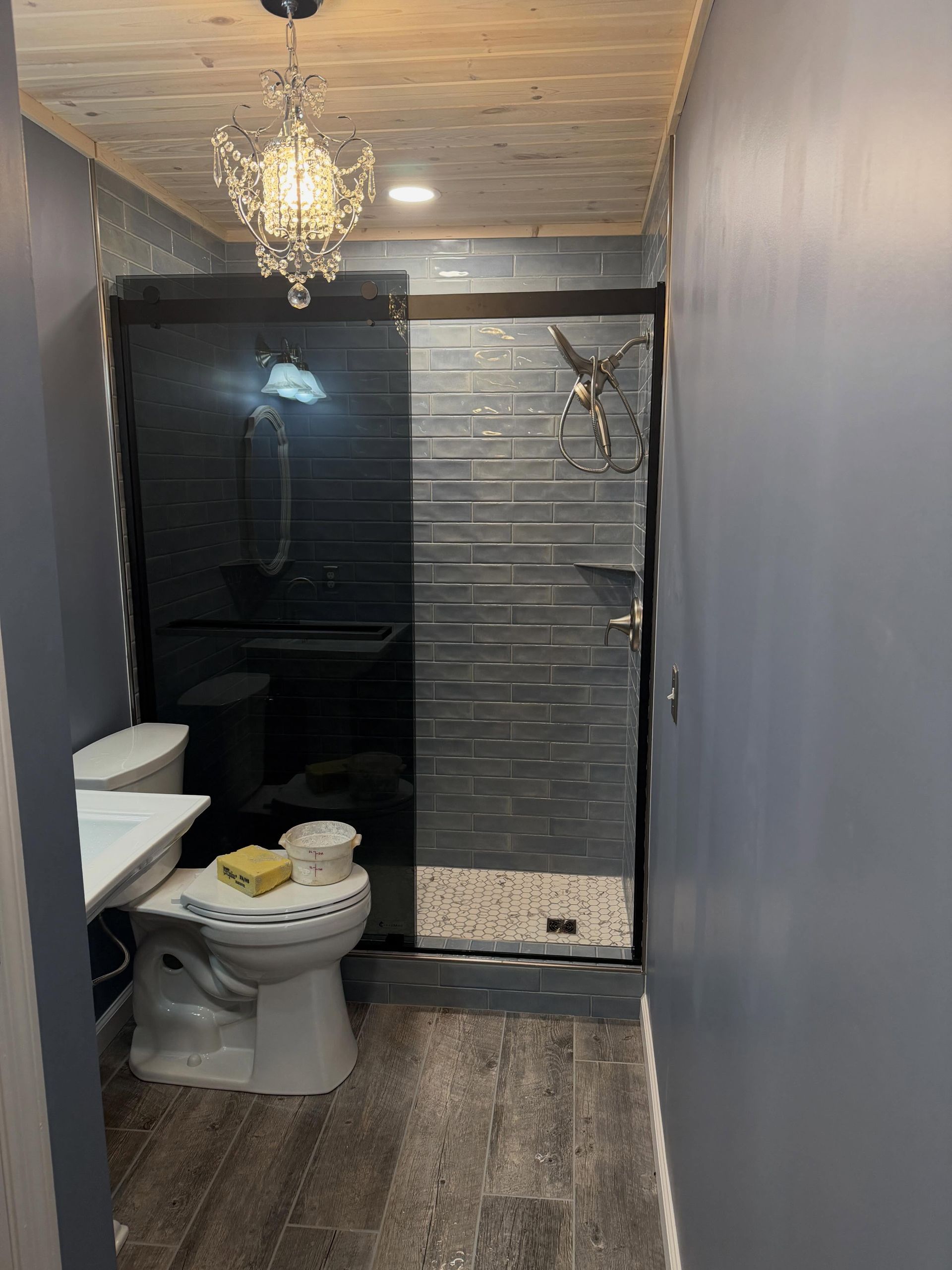 Small bathroom with toilet, shower, dark tile, gray walls, and a chandelier light fixture