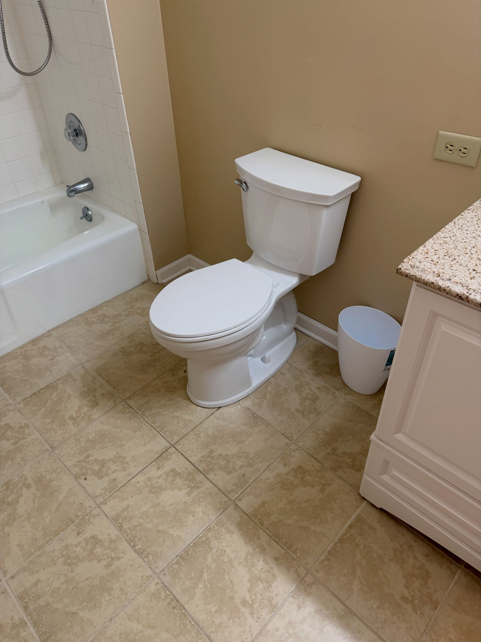 White toilet beside a bathtub and small trash can in a beige-tiled bathroom