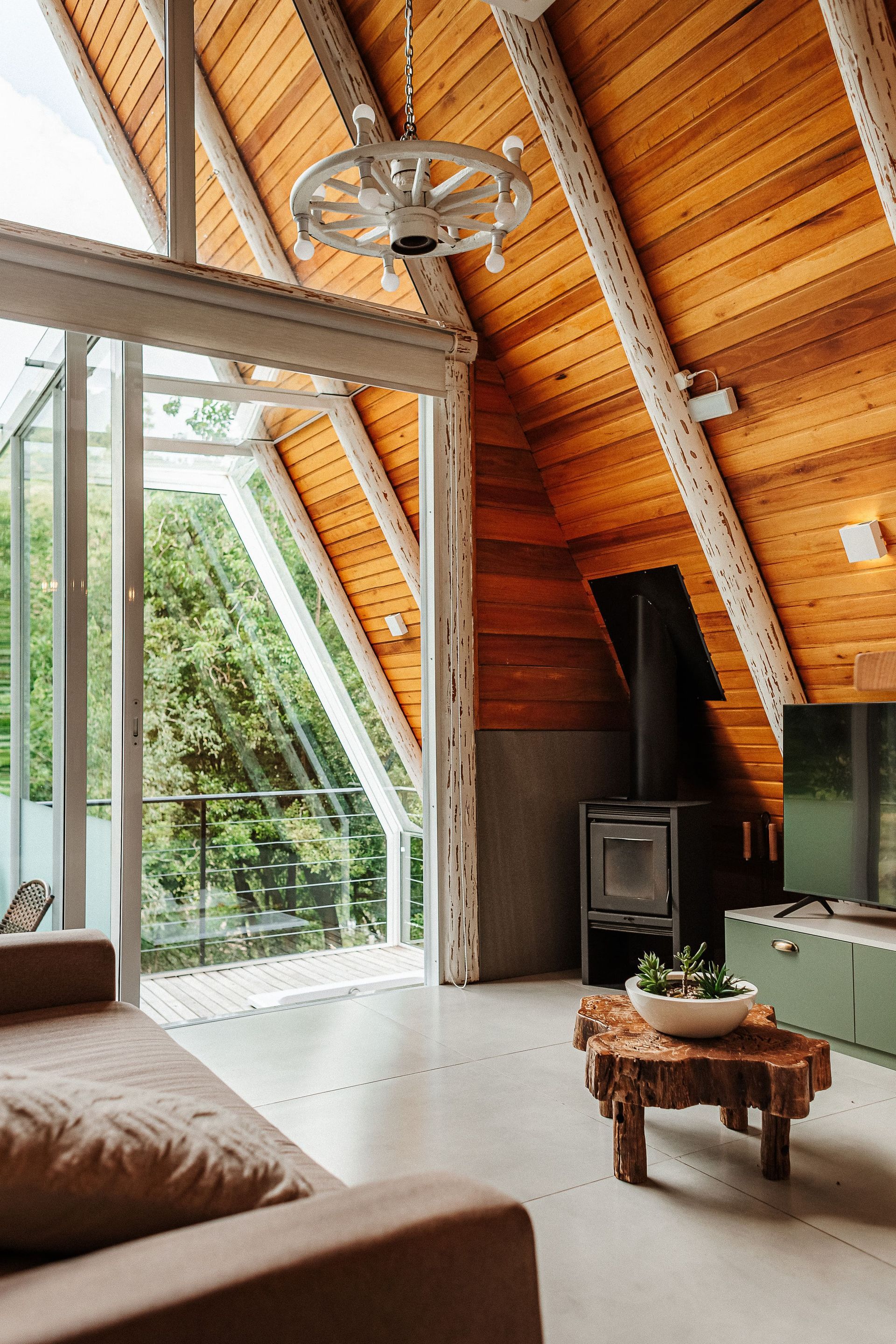 Sala de estar em estilo chalé com teto de madeira, janela ampla com vista para as árvores, sofá, mesa de centro e lareira.