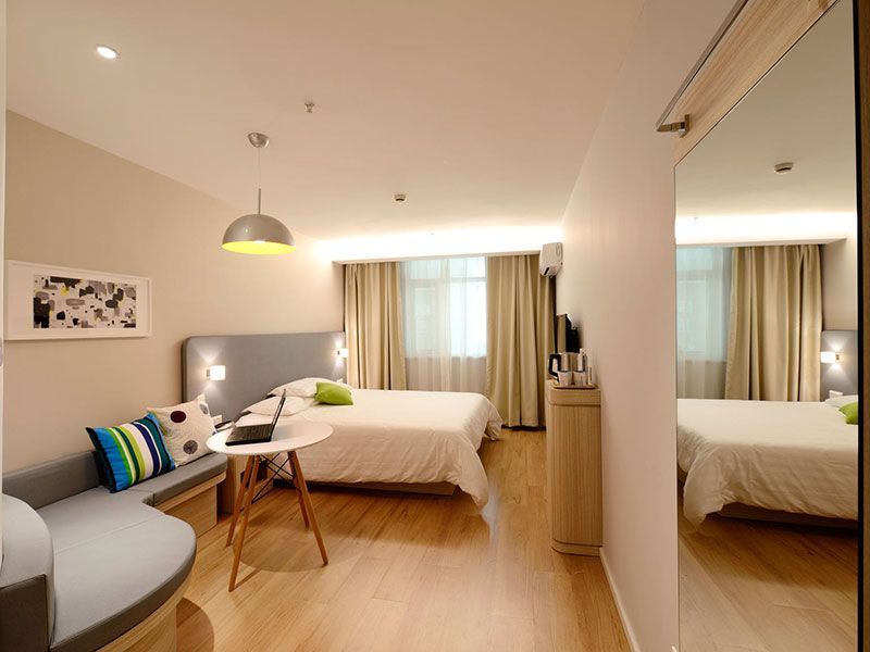 Quarto de hotel moderno com cama, sofá pequeno, mesa e espelho. Paredes bege e piso de madeira.