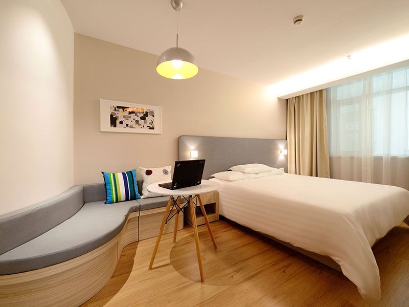 Quarto com cama, pequena mesa redonda com um laptop e um banco embutido curvo. Paredes bege e piso de madeira.