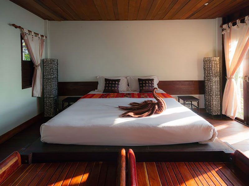 Quarto com cama grande, toalhas decorativas e detalhes em madeira. A luz do sol entra pela direita.