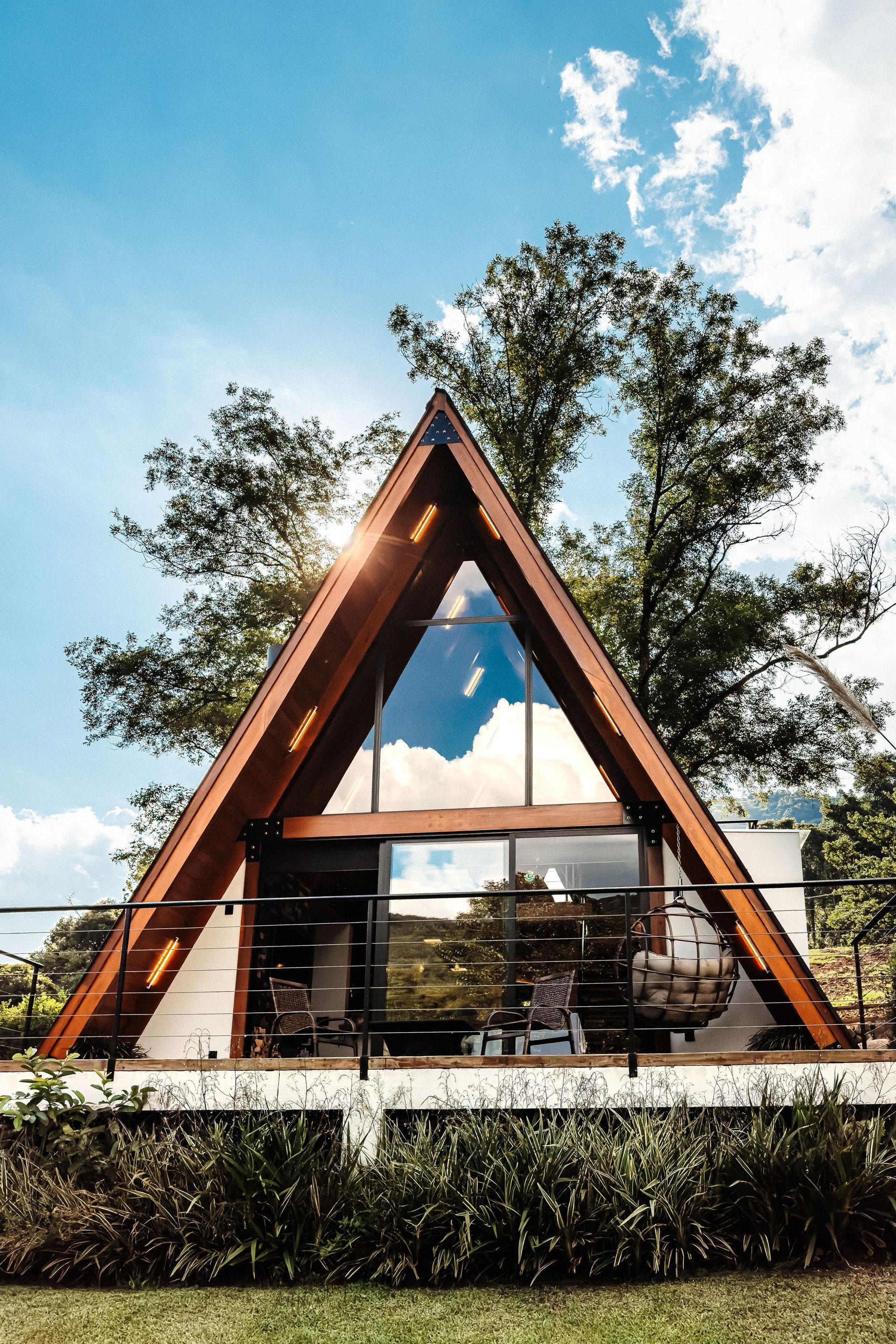 Cabana em forma de A com revestimento de madeira, grandes janelas de vidro e uma cadeira suspensa em um deck; sol brilhando através das árvores.