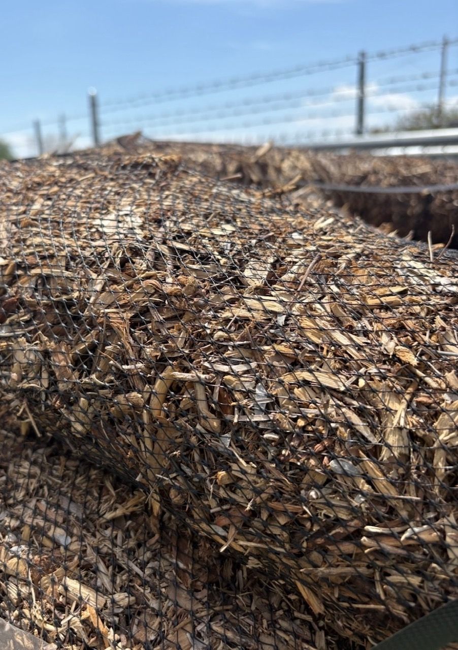 Pile of cut wood chips in a field, with a fence and blue sky in the background