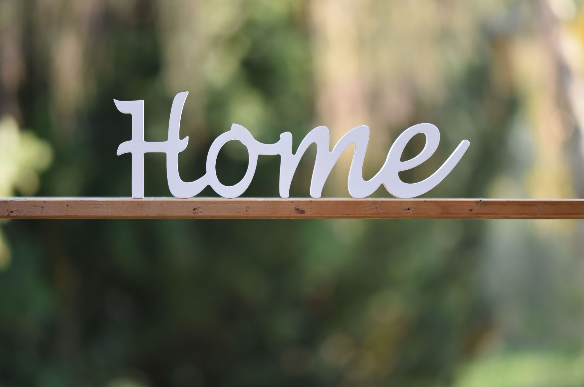 White cursive “Home” sign on a wooden shelf with a blurred outdoor background