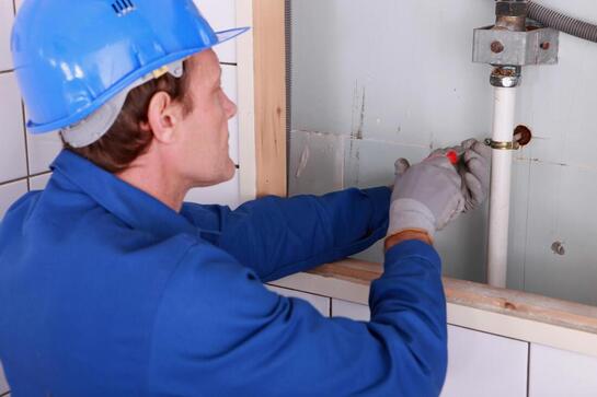 Plumber in blue uniform and hard hat repairing a wall-mounted pipe connection
