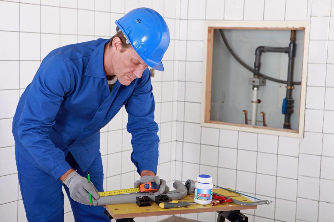 Worker in blue safety gear measures a countertop in a tiled room with exposed plumbing.