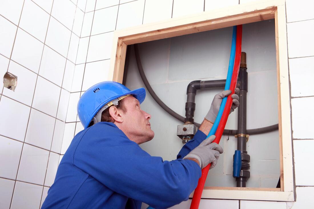Worker in blue hard hat installing plumbing inside a tiled wall opening with red and blue pipes.