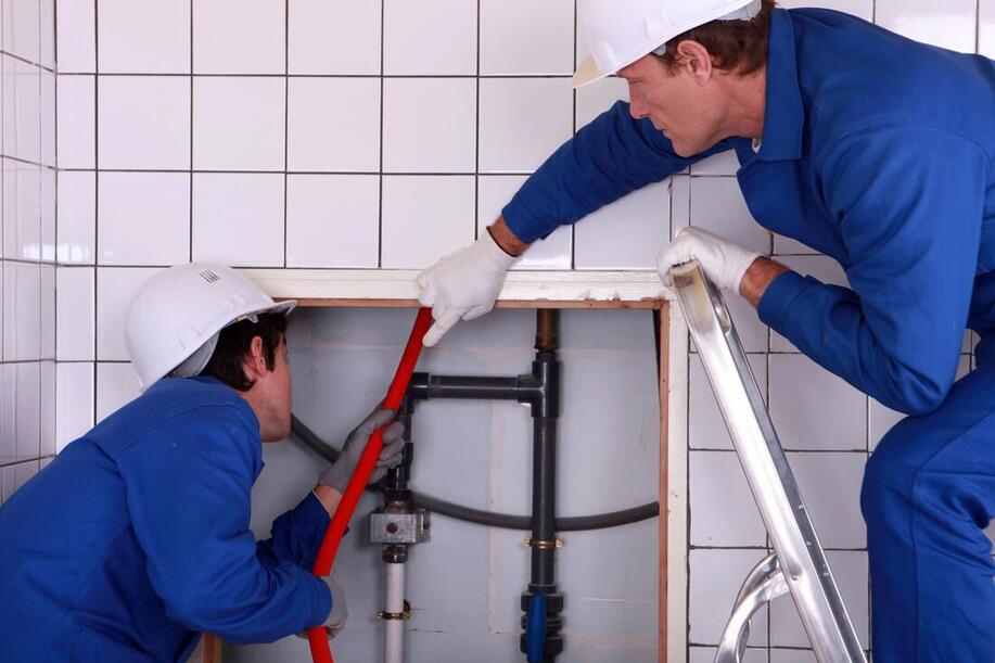 Two plumbers in blue uniforms repairing pipes in a tiled wall opening