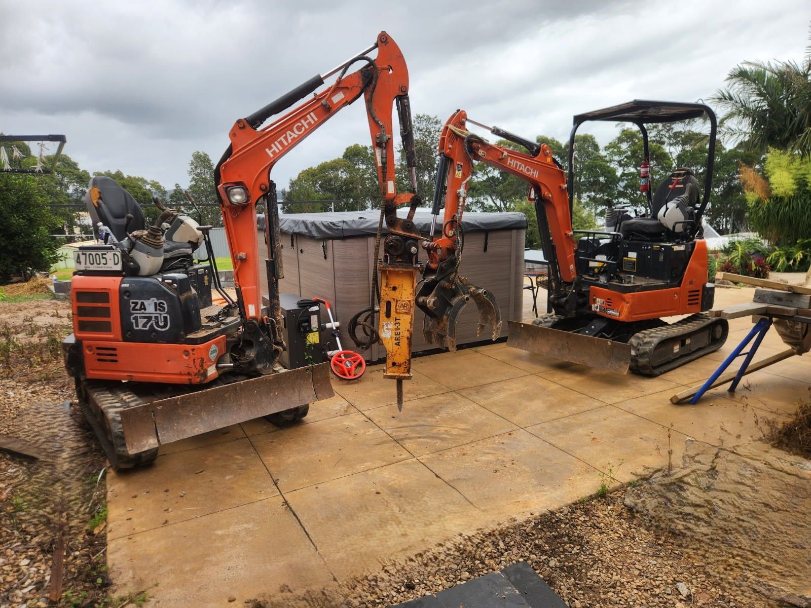 Assorted excavator and tractor buckets and augers arranged on gravel outdoors near rocks and plants