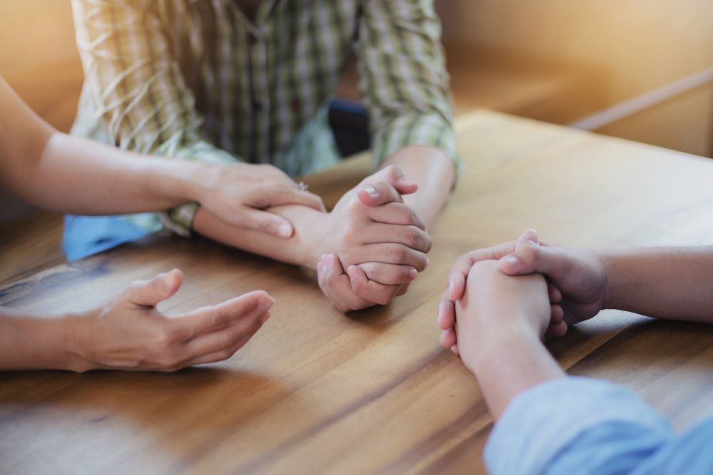 A Group Of People Are Sitting At A Table Holding Hands — Comfort N Care Support QLD Pty Ltd In Goondiwindi, QLD