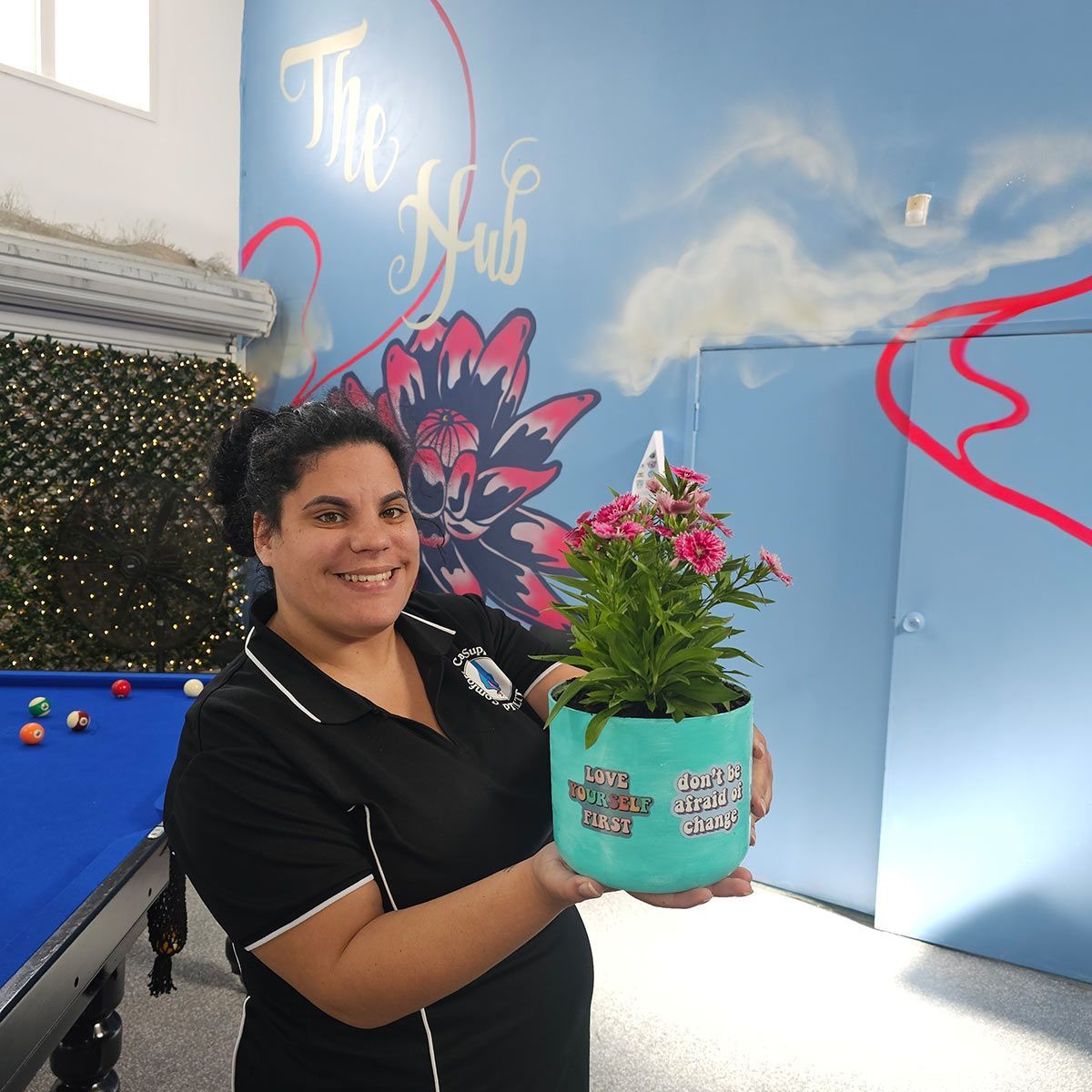 A Woman Is Holding a Potted Plant in Front of A Pool Table that Says the Hub — Comfort N Care Support QLD Pty Ltd In Toowoomba City, QLD