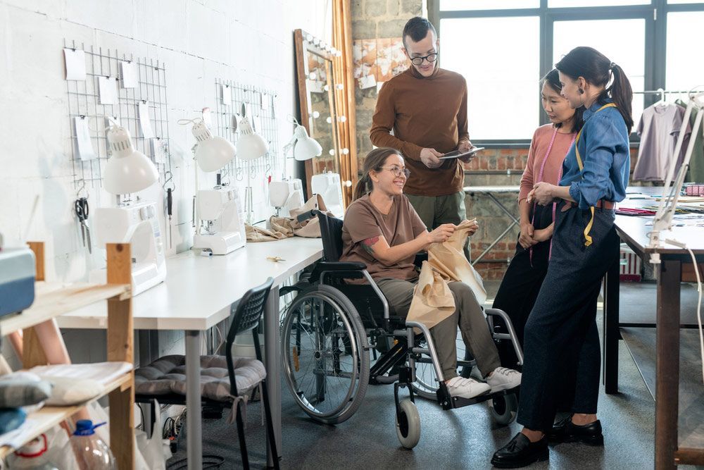 Group of young fashion Designers Surrounding Disable Seamstress In Wheelchair During Discussion — Comfort N Care Support QLD Pty Ltd In Toowoomba City, QLD