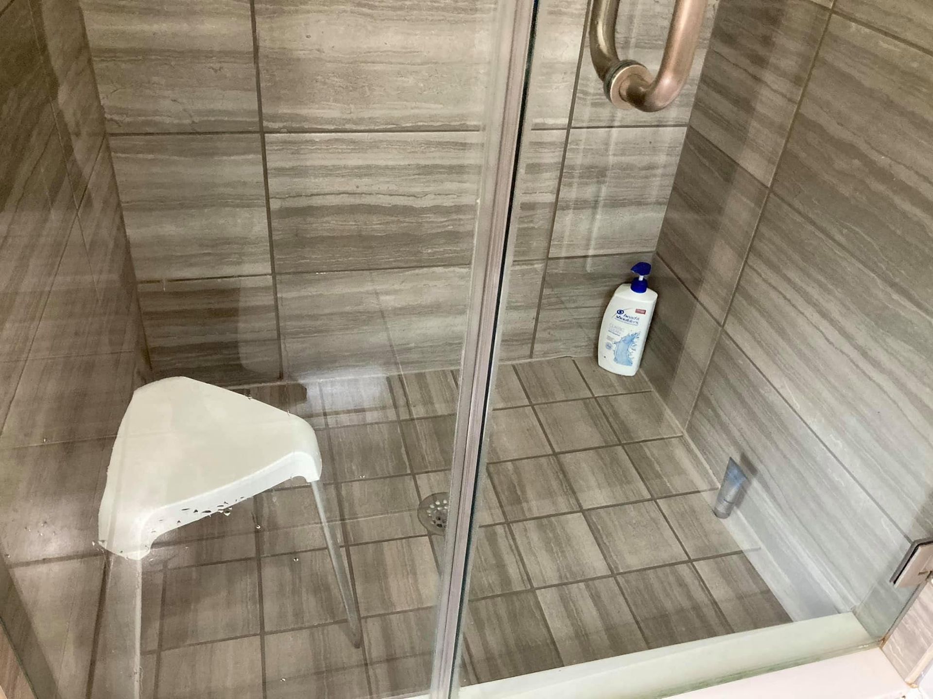 Shower stall with tiled walls, glass door, foldable white seat, and a bottle on the floor.