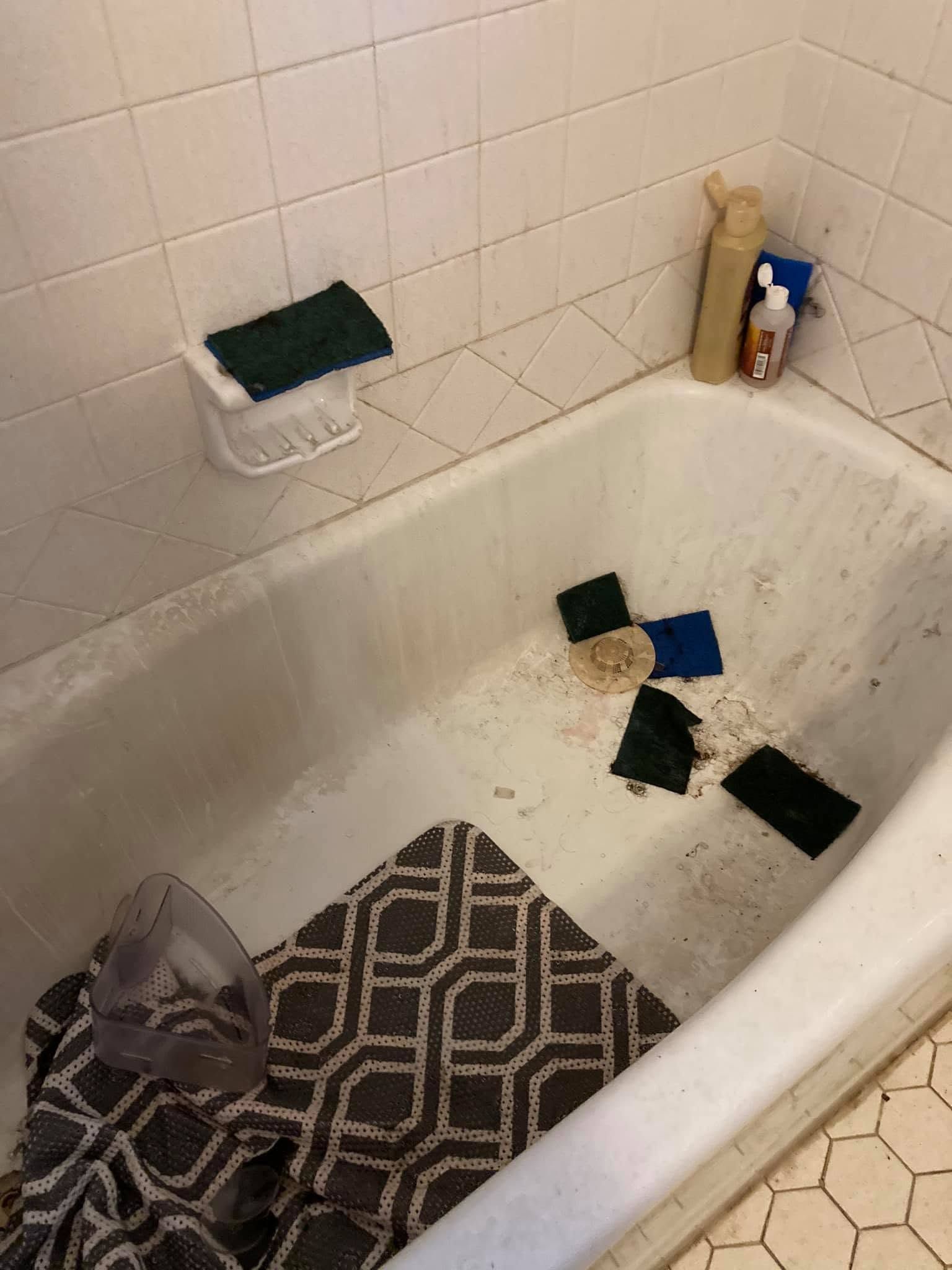 Dirty bathtub with scattered cleaning sponges and a bottle on the edge; bath mat and trash bag nearby