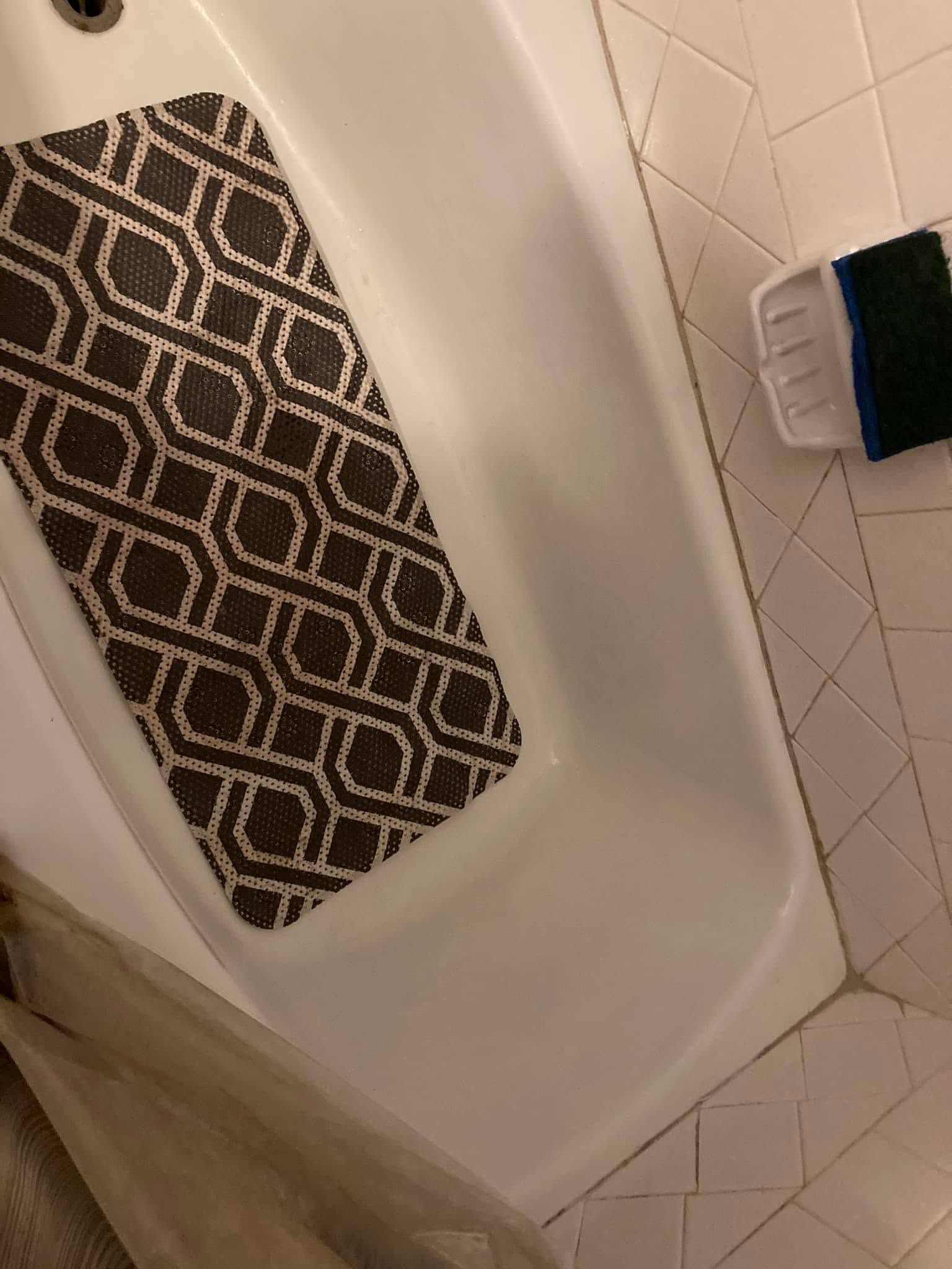 Bathtub with a brown patterned mat and a blue-handled brush on the tiled wall