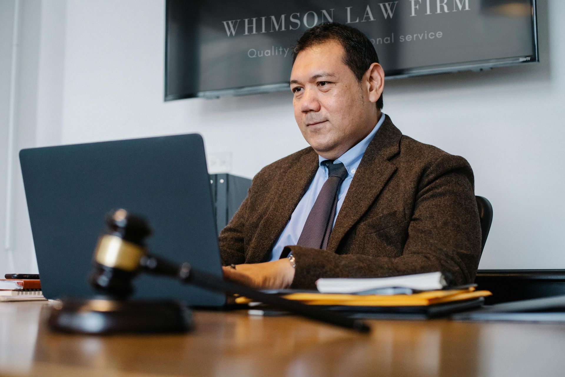Man in a brown blazer sitting at a desk in an office, with a laptop and legal documents in front of him.