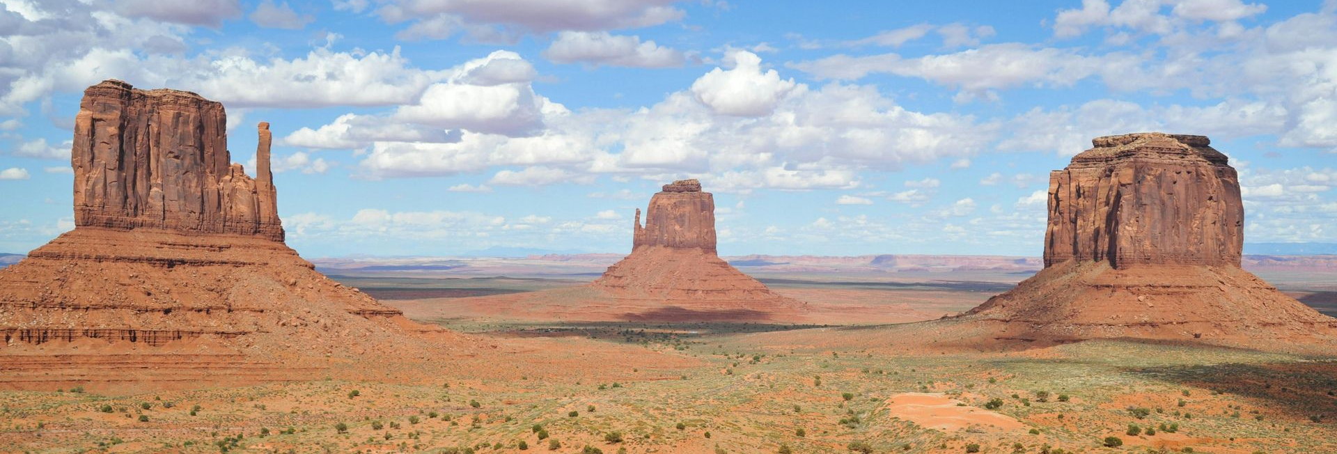 Desert landscape with three red sandstone buttes under a partly cloudy blue sky.