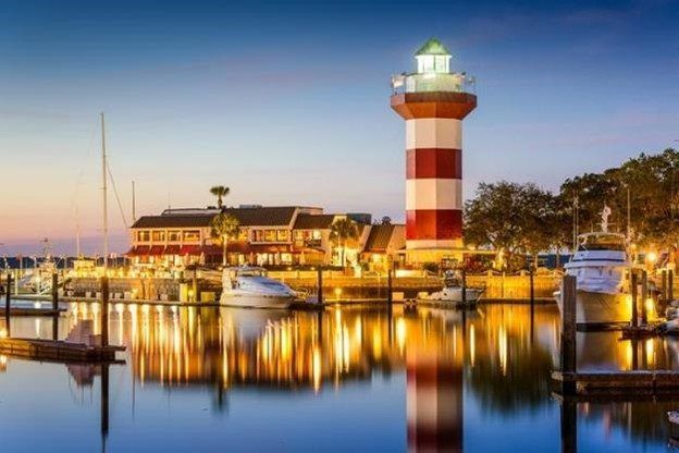 Harbor at dusk with a red-and-white lighthouse, boats, and glowing waterfront buildings reflected in calm water