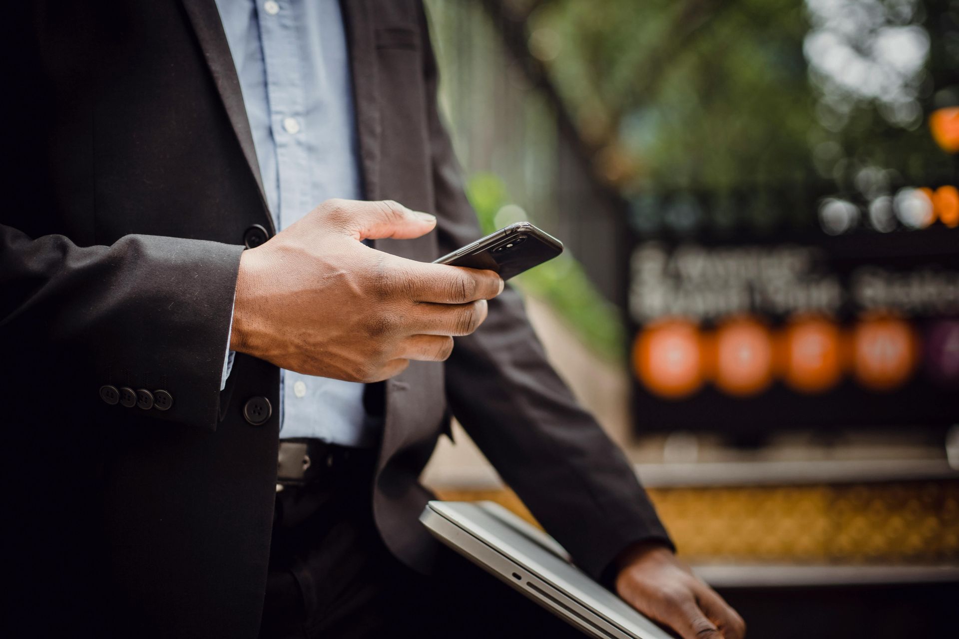 Man in dark suit holding a smartphone and a folder outdoors near a blurred fence and lights